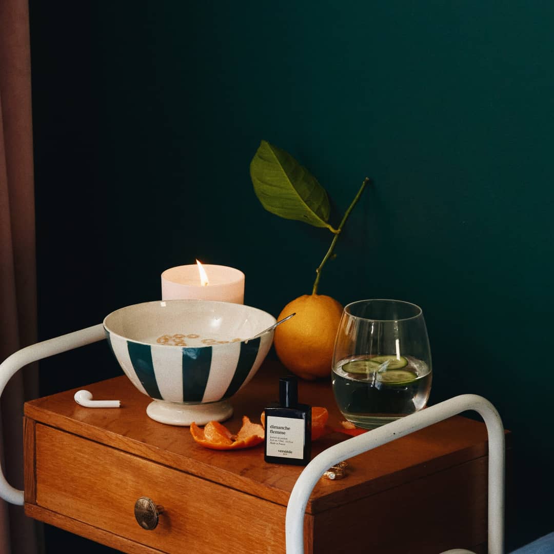 Flacon Dimanche Flemme de Versatile Paris sur une table de chevet avec bougie et orange